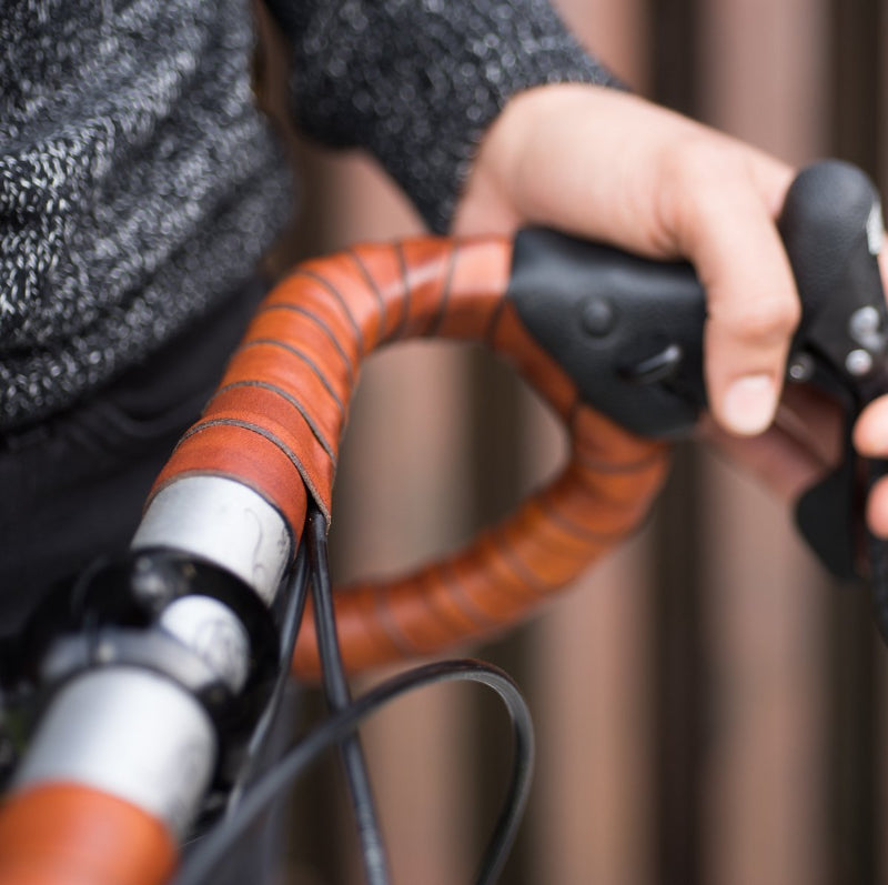 Close-up of bicycle drop bar with honey leather straps wrapped in neat coils on the drop bar. A Black woman cyclist's hand is on the left-hand brake lever.