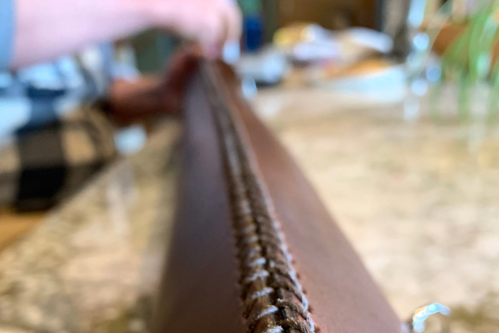 Close-up of a dark brown leather tube being hand-stitched on a marble kitchen countertop, with Geoff in the background stitching it with a needle using his hands.
