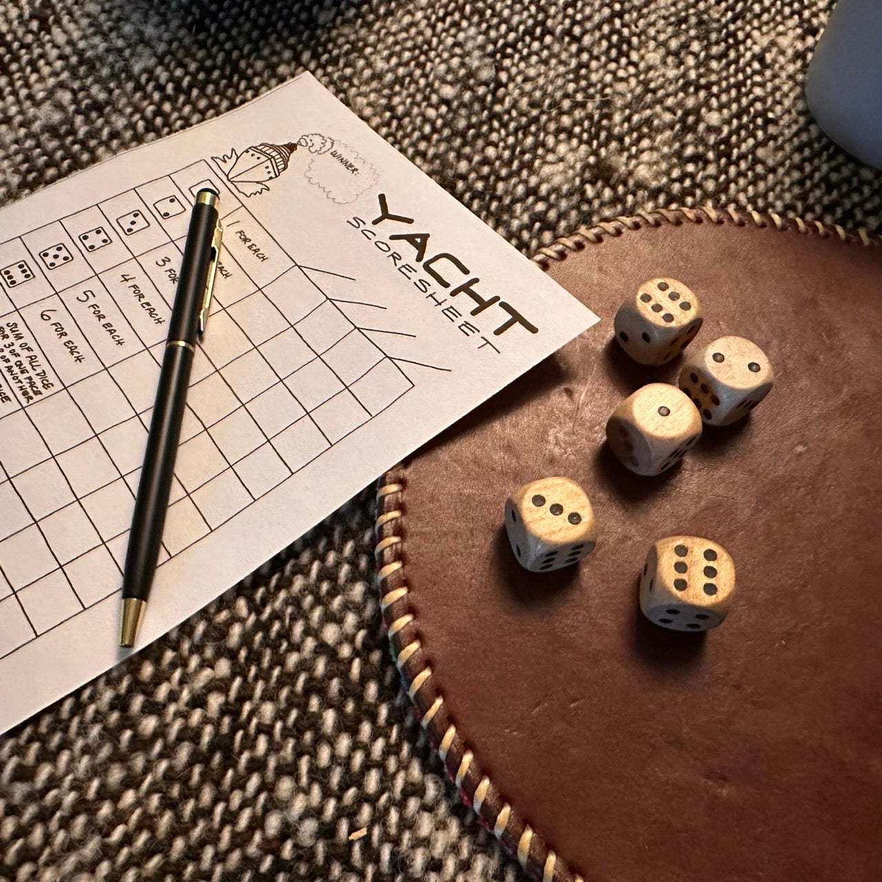 Wooden dice on a brown leather surface with a sheet of paper that says Yacht Scoresheet and a pen