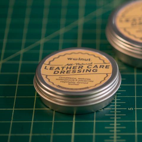 Two tins of Walnut Studiolo All Natural leather care dressing on a green grid background, label says, Conditions, restores, waterproofs and preserves vegetable tanned leather