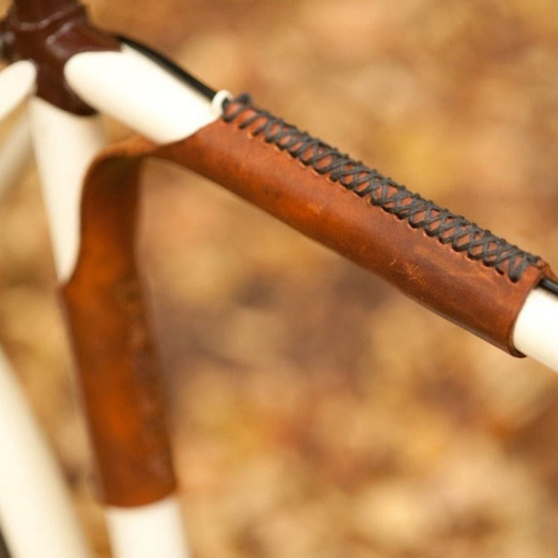 Close-up of a bicycle frame near the seat tube with a brown leather sew-on strap that bridges the top tube and the seat tube against a blurred natural background of fallen leaves