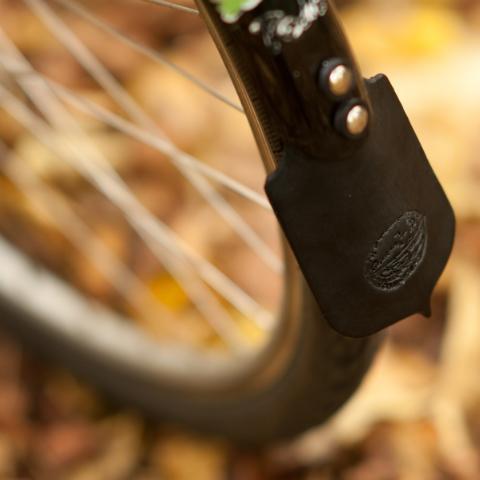 Close-up of a bicycle wheel with a black fender and leather mud flap against a background of blurred autumn leaves