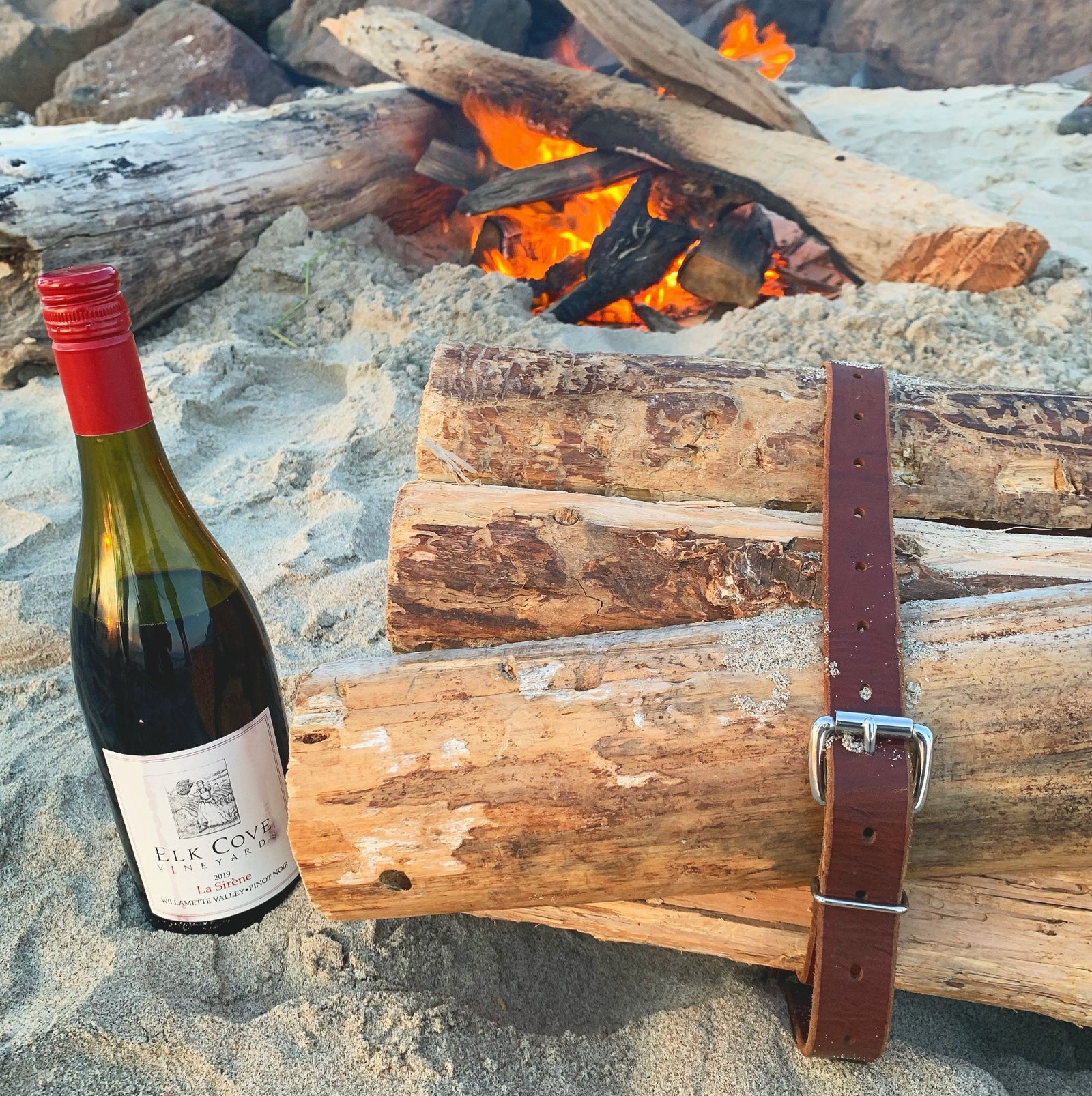 Wine bottle on the beach next to a bundle of firewood held together with a brown leather belt belt carrier with a fire in the background