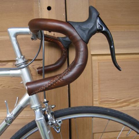 Dark Brown sew-on leather bar wraps on a set of bicycle handlebars (drop bars). This is a close up profile view of the handlebars against a raw wood backdrop.