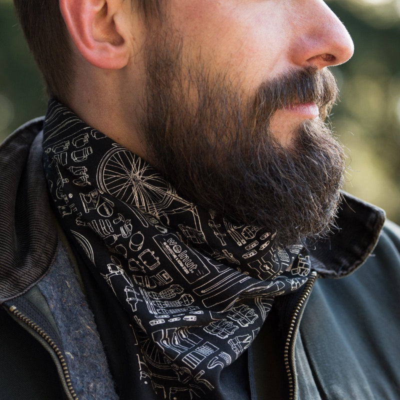 Beared man wearing a bicycle patterned bandana on his neck with a black canvas jacket with a blurred natural background