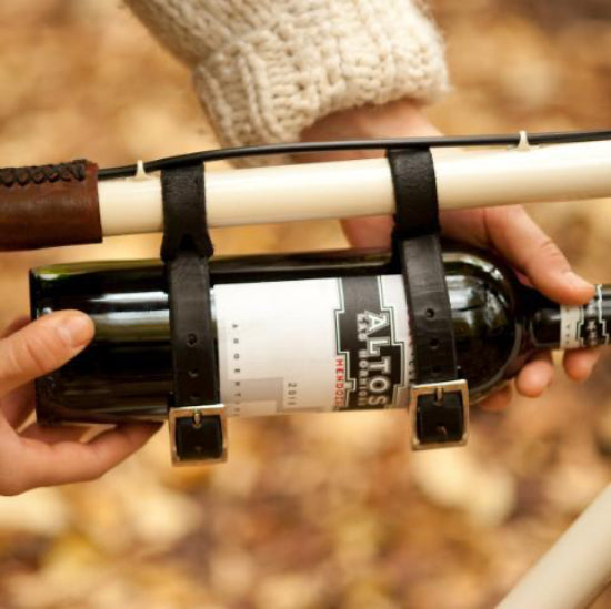 Hands touching a wine bottle that is mounted with leather straps to the top tube of a cream colored bicycle on a fall today in front of background of fallen autumn leaves