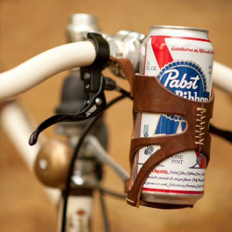 A can of tallboy PBR in a leather hand-stitched bicycle cage mounted to the handlebars of a bicycle