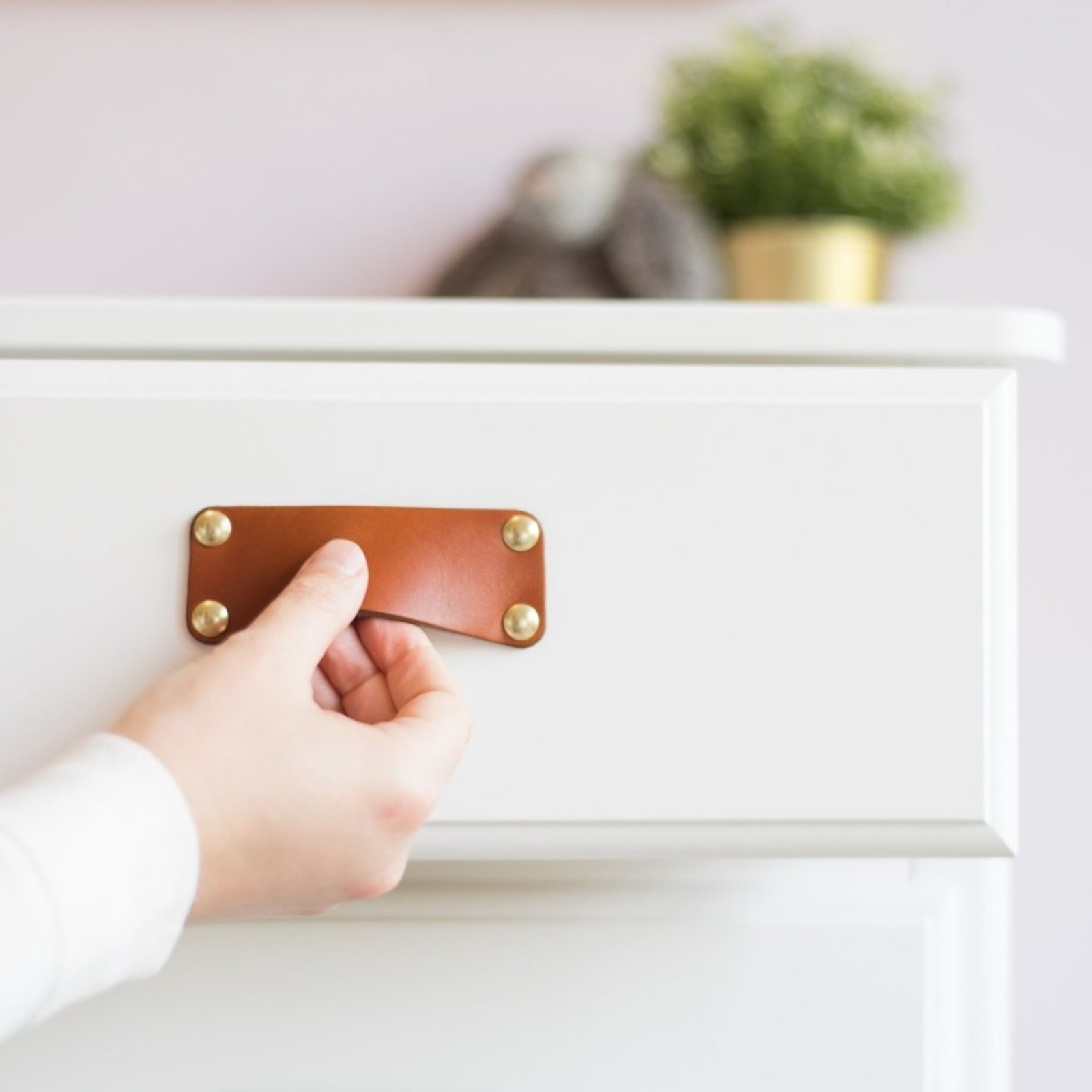 Woman's hand is opening a drawer in a white chest of drawers using a honey leather bin pull with brass hardware.