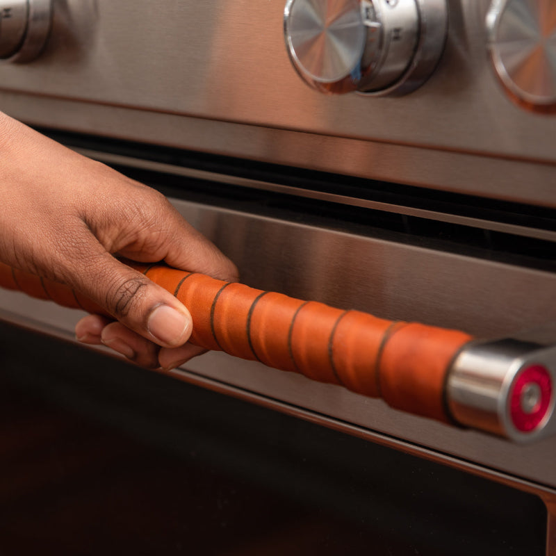 Close-up of a woman's hand opening a stainless steel oven door that is wrapped in honey vegetable tanned leather