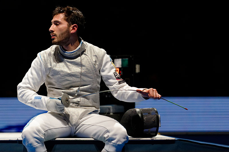 ﻿A young Italian man sitting sweaty in an Olympic stadium, wearing a full white protective fencing outfit and wiping his foil fencing blade