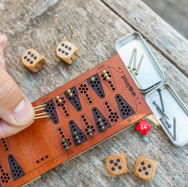 Close-up view of a small leather travel backgammon board with brass and black game pieces and parts outdoors on a weathered wood picnic table