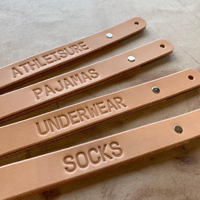 Four natural colored tan leather handles on a work surface embossed with the words, Athleisure, Pajamas, Underwear, Socks