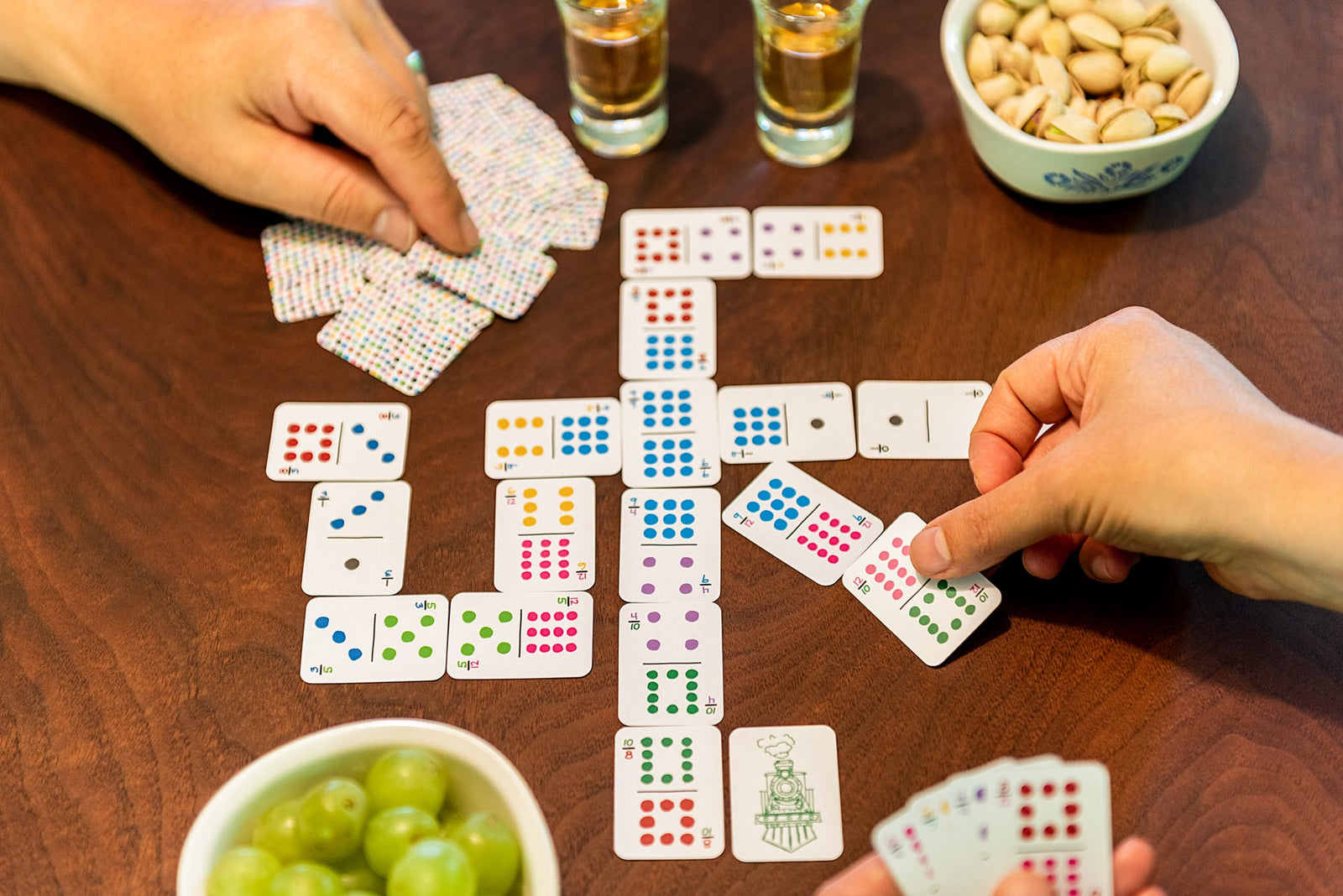 Two people playing dominoes with colorful domino playing cards on a walnut wood table with snacks and drinks
