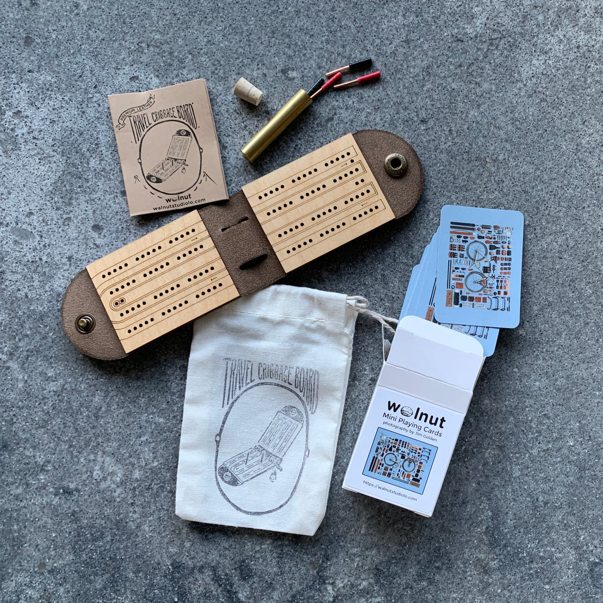 Open leather and wood travel cribbage board with wire metal pegs on a tabletop with a carrying pouch and a half-sized deck of cards, which is the "Ultralight Gift Set"