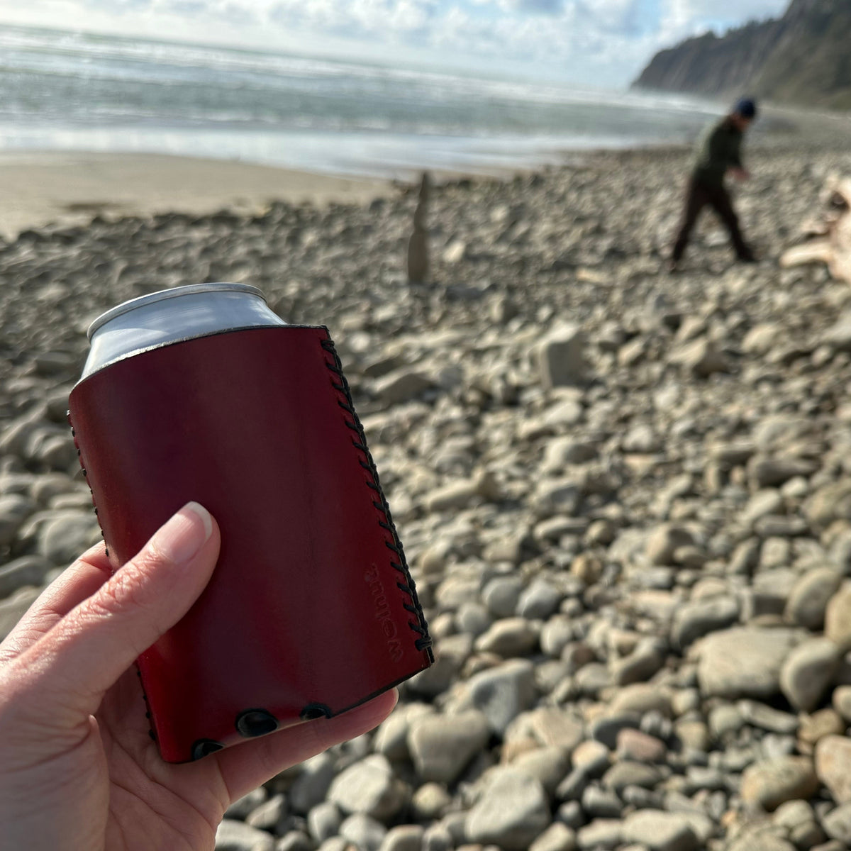 Hand holding a can with a red leather koozie on a rocky beach with ocean waves and a person in the background.