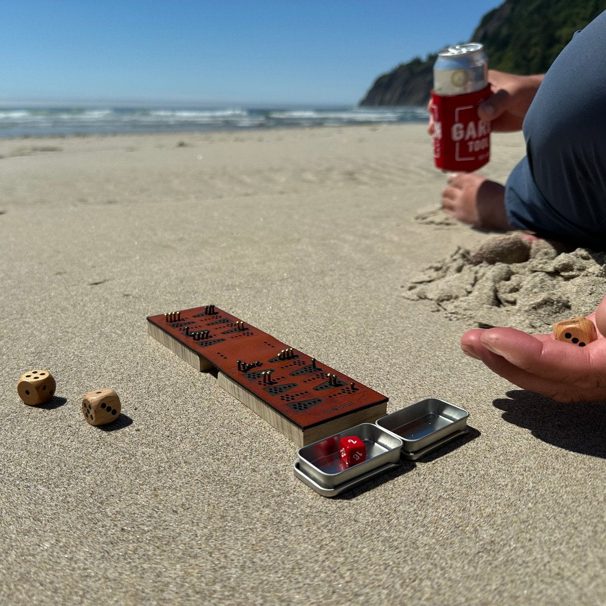 Photo taken at the beach, with a travel backgammon board on the sand, the camera facing the ocean. A person is sitting to the right side mostly off-camera, rolling dice in his hand.A relaxing way to play backgammon on the go - "beachgammon"?