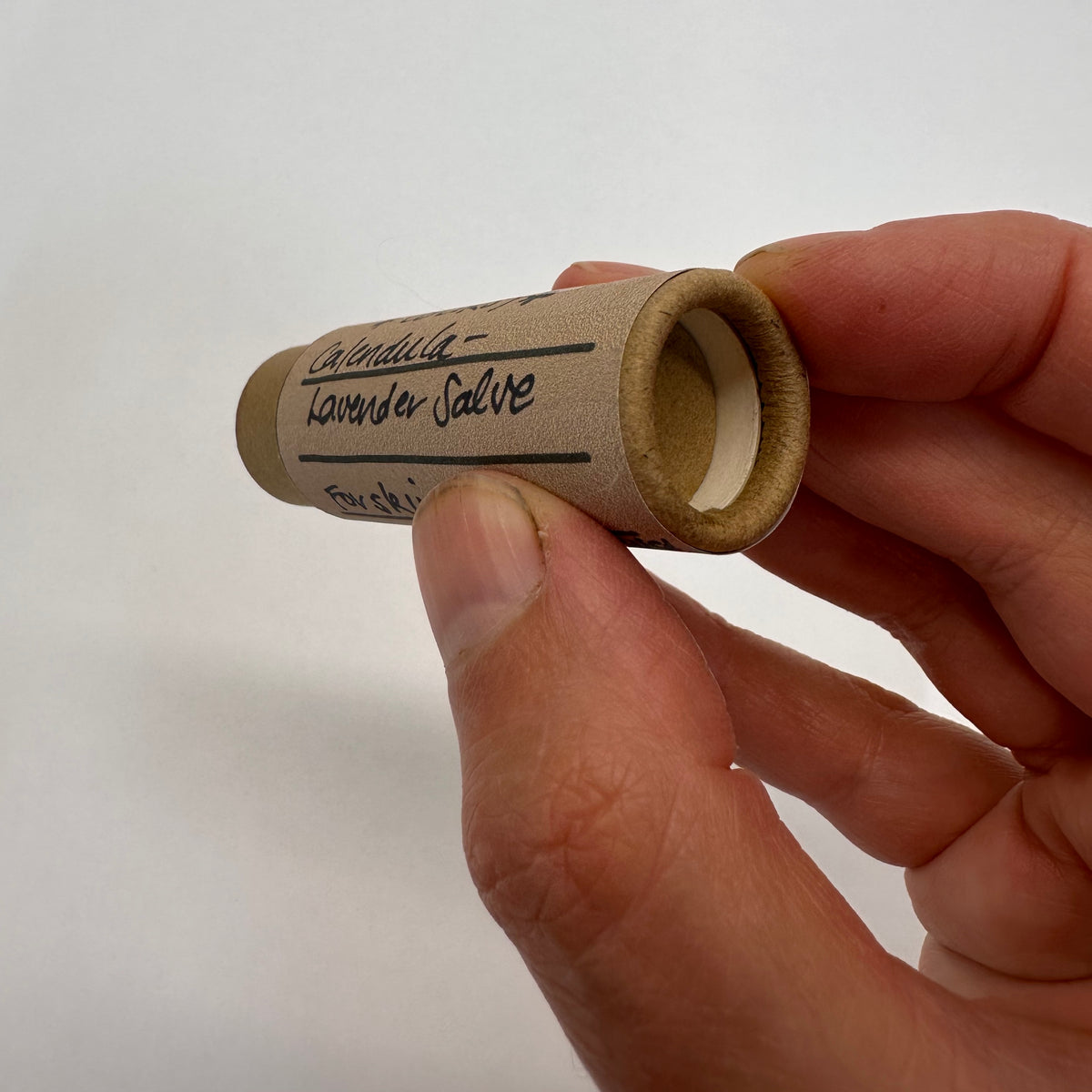 Hand holding a small cardboard tube labeled 'Lavender Salve' against a white background showing the bottom where the finger will push up