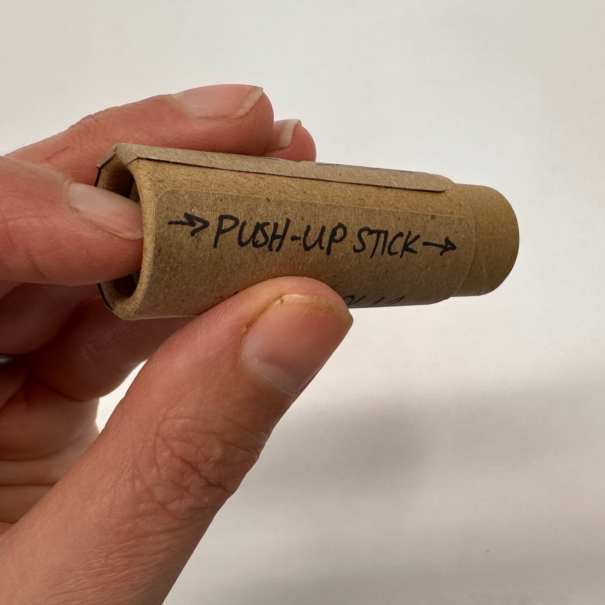Hand holding a cardboard tube labeled 'Push-up Stick' against a plain background with the finger pushing up to dispense