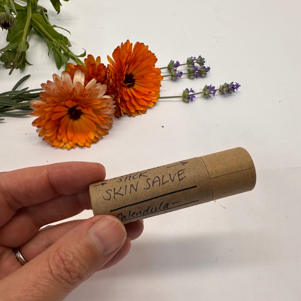Hand holding a skin salve stick labeled 'Calendula' with flowers in the background.