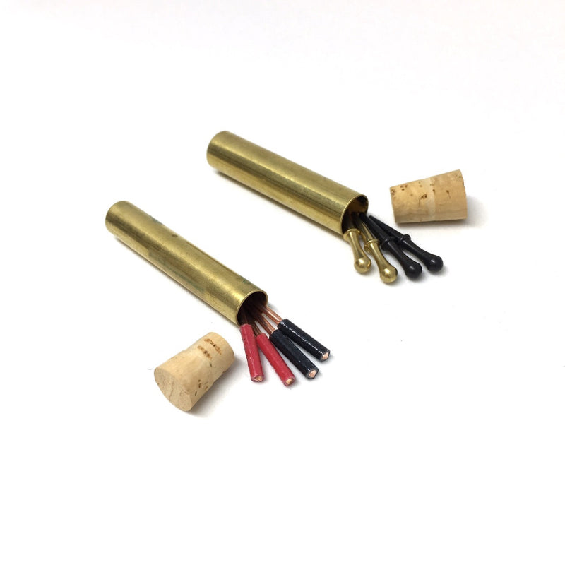 Two brass storage tubes with cork closure ends and small travel mini cribbage pegs spilling out in two styles: colorful wires and cast pegs, on a white background