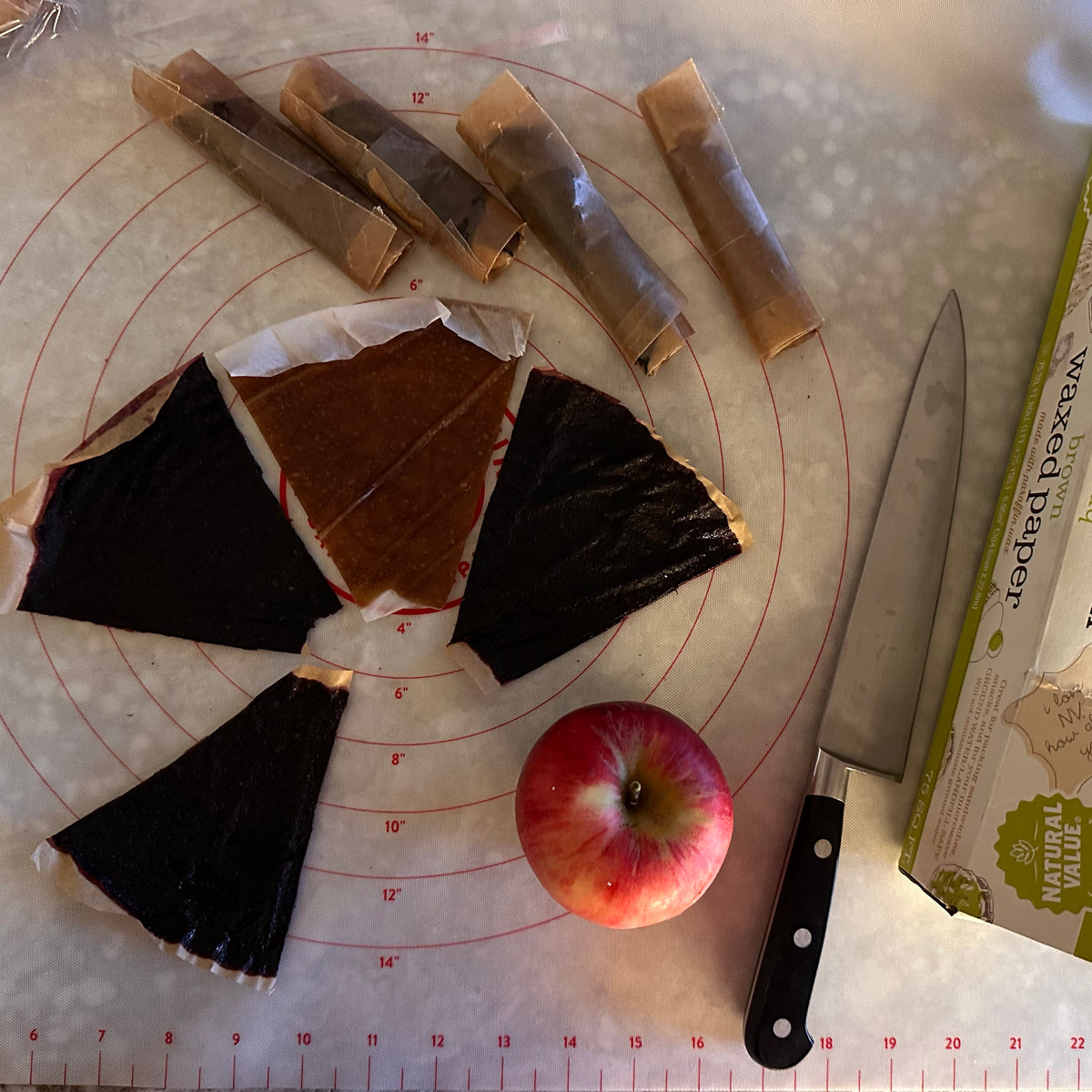 Cutting mat from a bird's eye view, with wax paper wrapped fruit rolls, cut fruit rolls that are about 1/8 of a dehydrator tray, an apple, and a knife