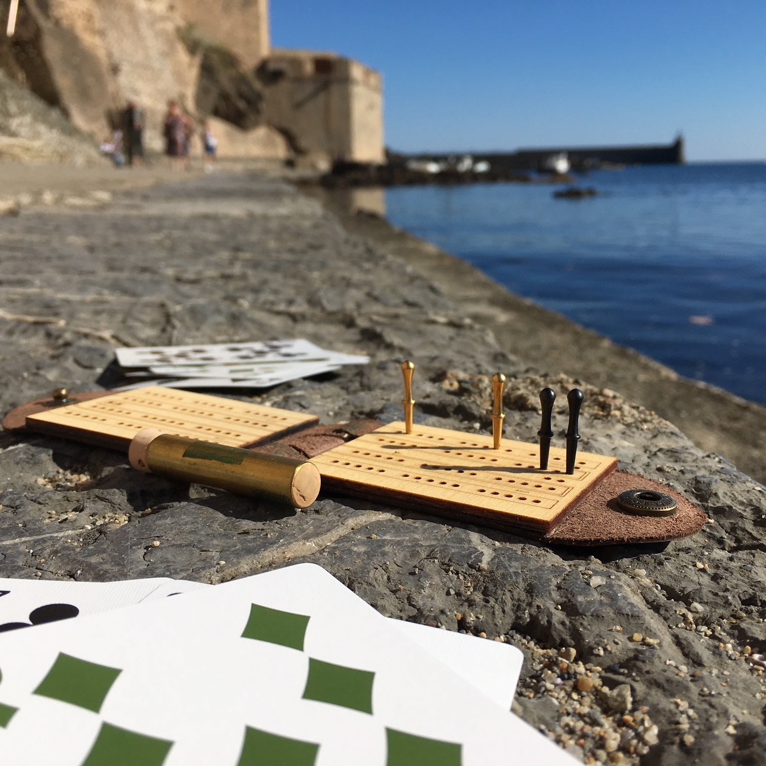 A leather travel cribbage board set up on the walls of a causeway in the Mediterranean, with a castle visible in the background, in Collioure France. 