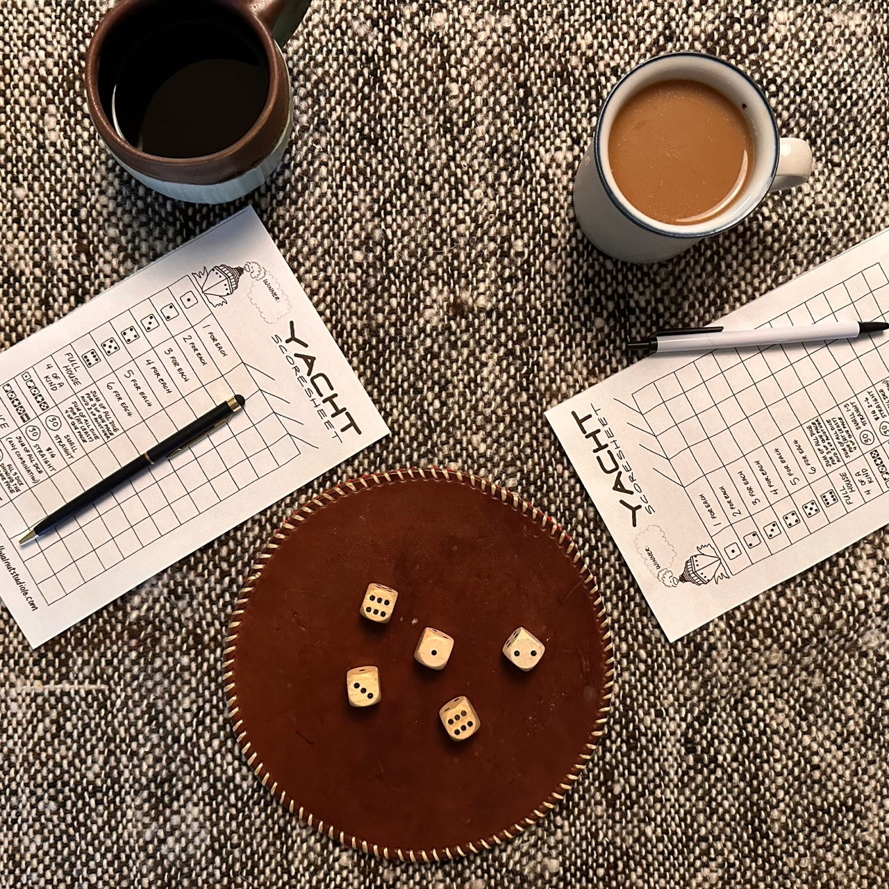 Yacht (Yatzy) game set-up on a coffee table including dice, coffee cups, and score sheets with pens on a textured surface.