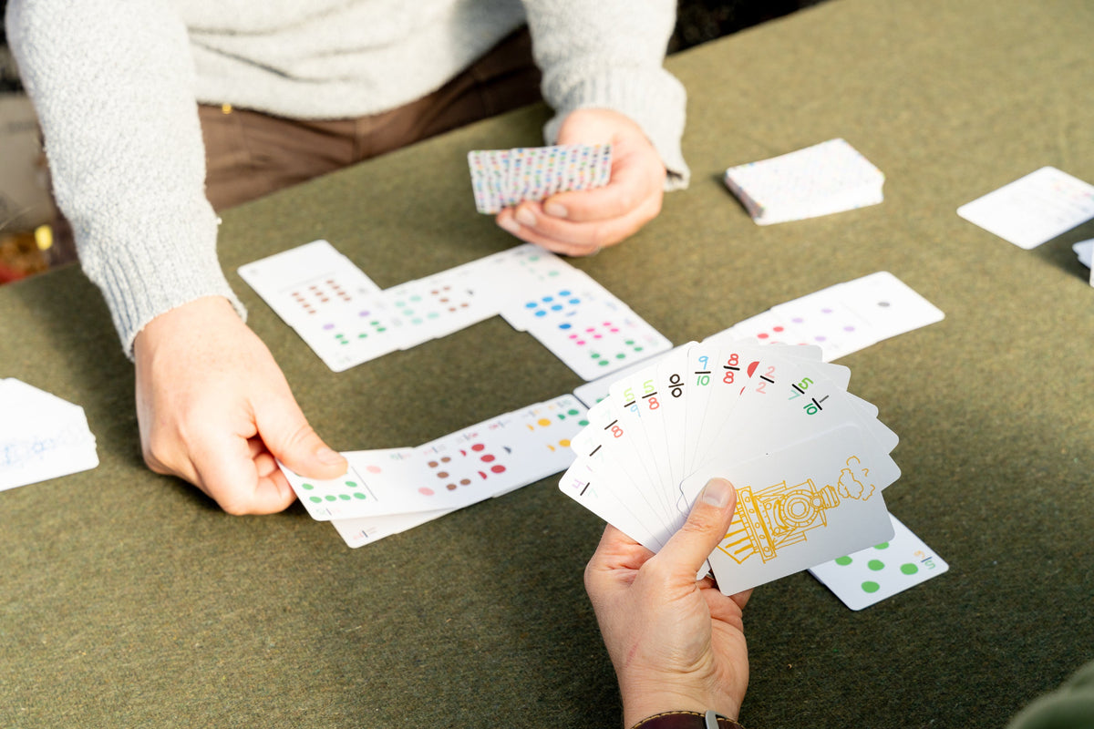 A close-up lifestyle shot of people playing a card game at a green table. Two hands are visible, one holding a fan of playing cards with a gold illustration and others showing colorful card faces featuring a Mexican Train token card. 