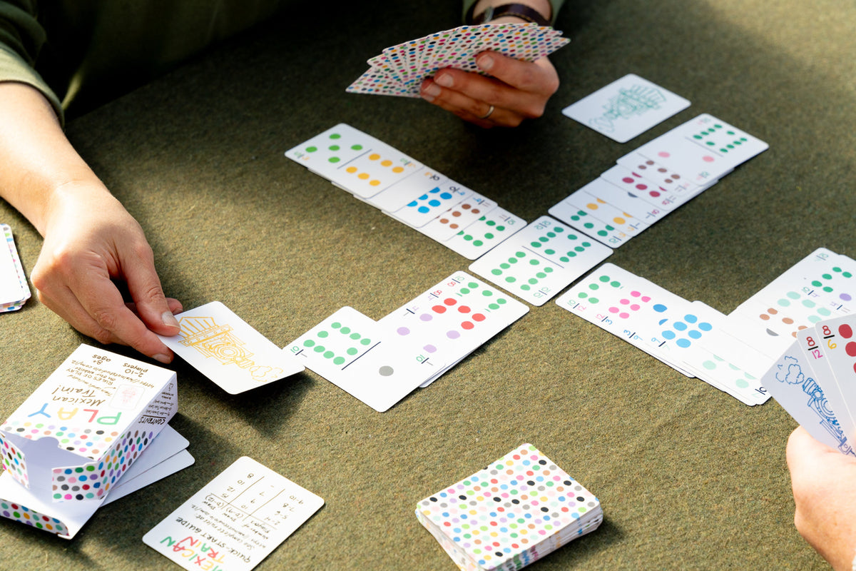 Several people are playing a card game at a table, with cards and card boxes spread across a dark green surface. The cards and boxes feature colorful polka dots and printed text.