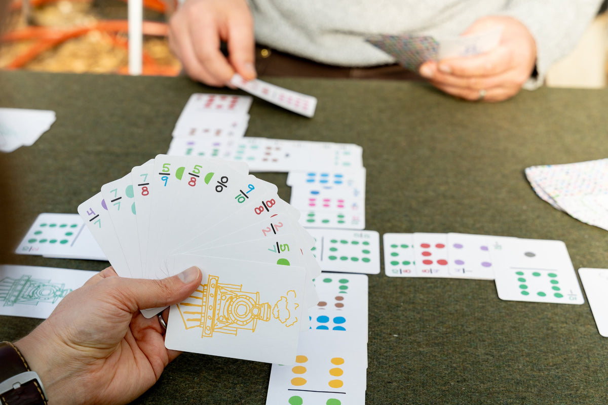 A close-up of a domino card game in progress on a dark table, with a hand holding a fan of playing cards in the foreground and another person's hands visible at the top placing a card onto the game board.