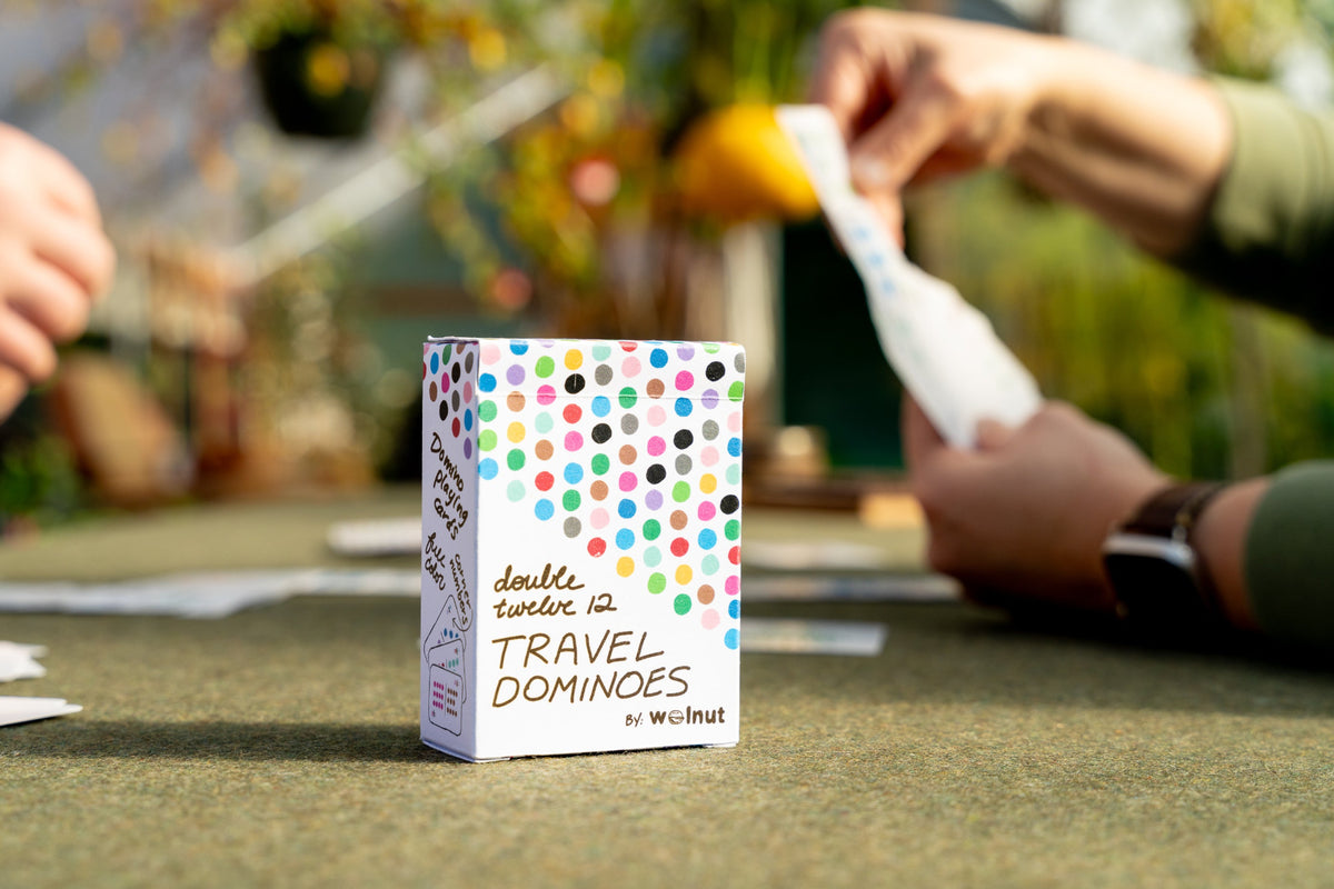 A boxed card game called "double twelve Travel Dominoes" stands on a table in an outdoor setting, with hands holding cards in the blurred background.