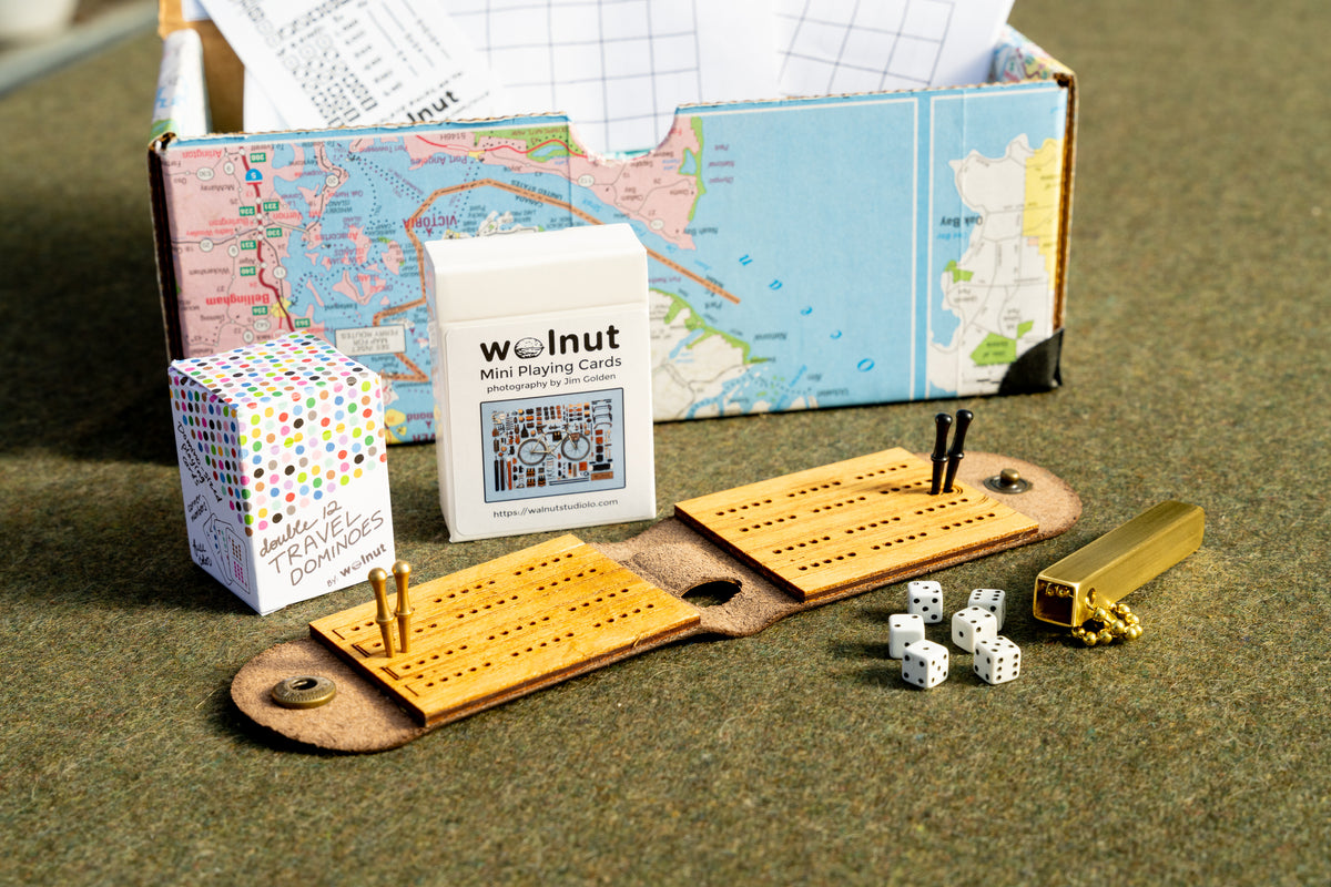 "backpacker's" Travel games gift bundle shown with a handcrafted memory box, "places I've been" keepsake map, printed scorecards for Mexican Train, Farkle / 10,000 and Yacht / Yatzy dice games, travel dice in a brass tube, leather cribbage board, half-size travel playing cards, leather backgammon board, and double 12 travel dominoes playing cards 