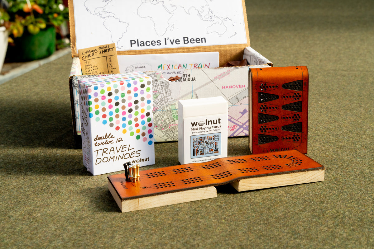 "Game Lovers" Travel games gift bundle shown with a handcrafted memory box, "places I've been" keepsake map, printed scorecards for Mexican Train, and cribbage counting cheatsheet, leather cribbage board, half-size travel playing cards, leather backgammon board, and double 12 travel dominoes playing cards 