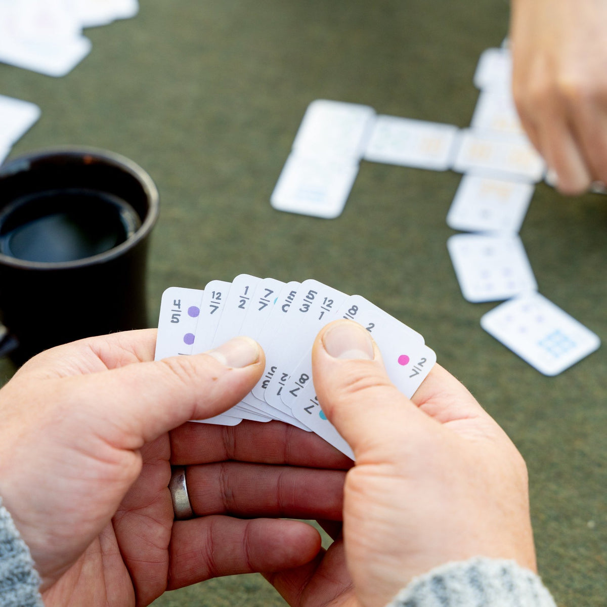 Looking over the shoulder of a person playing Mexican Train domino game using Double 12 domino playing cards. She is holding the cards in her left hand and picking one out with her right to place on the table. 
