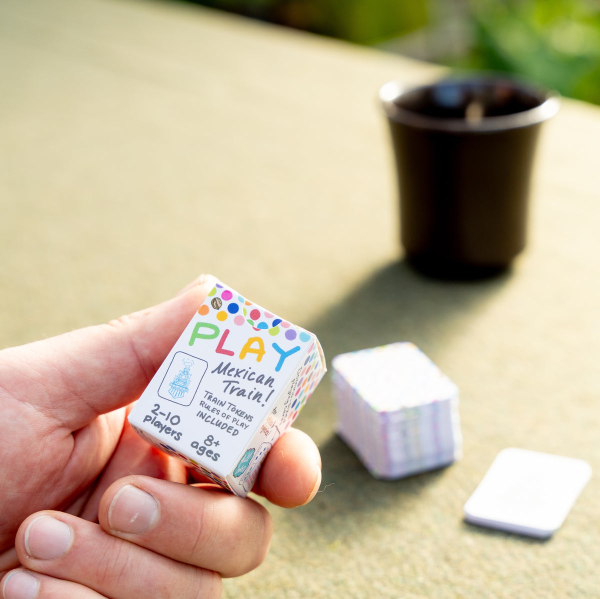 A man's hand holding the small box of travel domino playing cards between his thumb and forefinger. Blurred out in the background is a sunny table with a green tablecloth, the stack of cards ,and a cup of coffee. On the box, he's reading, Play Mexican Train! 2-10 Players, Ages 8+