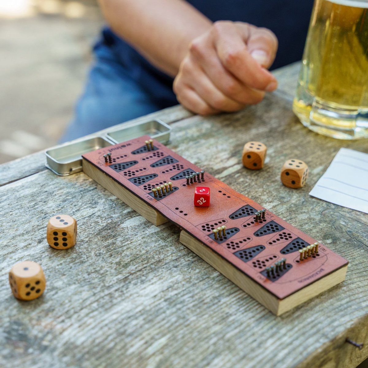Close-up of a leather travel backgammon travel board opened and set up for play on a picnic bench outdoors. A person's hand is visible in the background of the photo along with a beer. Fun times! 