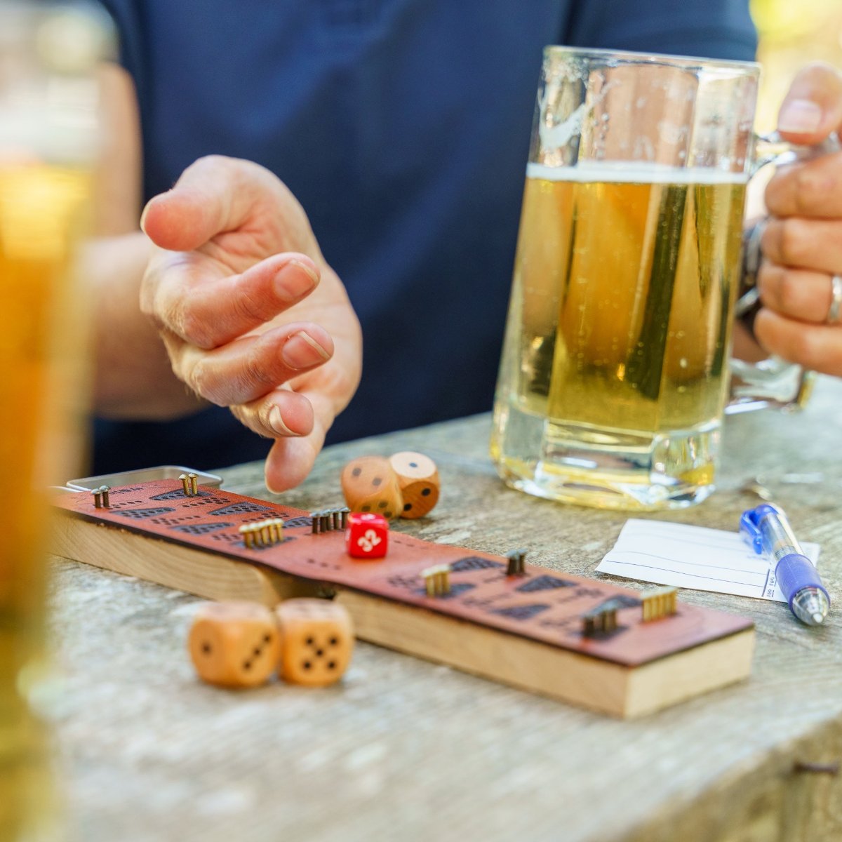 A person wearing a navy blue shirt is rolling wood dice on a picnic table next to a leather and wood travel backgammon board. They are rolling with their right hand and the dice are tumbling down from their hand inmotion, and in their left hand they're holding a stein of golden yellow beer. 