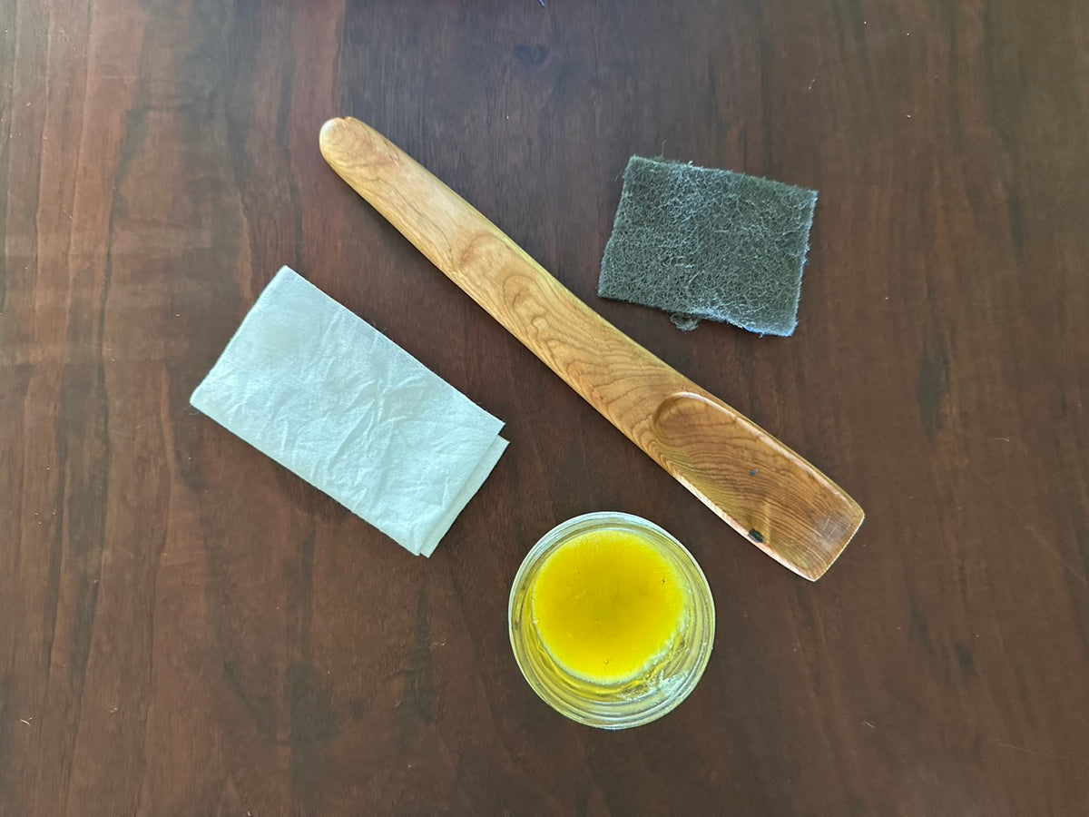 Still-life of a hand-carved wood spoon, abrasive hand pad, cotton buff rag, and flax wax in a rich gold color