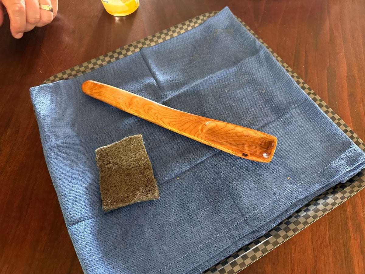 A wood spoon with a shiny glare resting on top of a blue cloth with an abrasive pad next to it