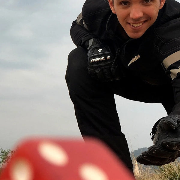 Person in black motorcycle gear posing outdoors with a cloudy sky and rolling a large red dice to the camera in the foreground
