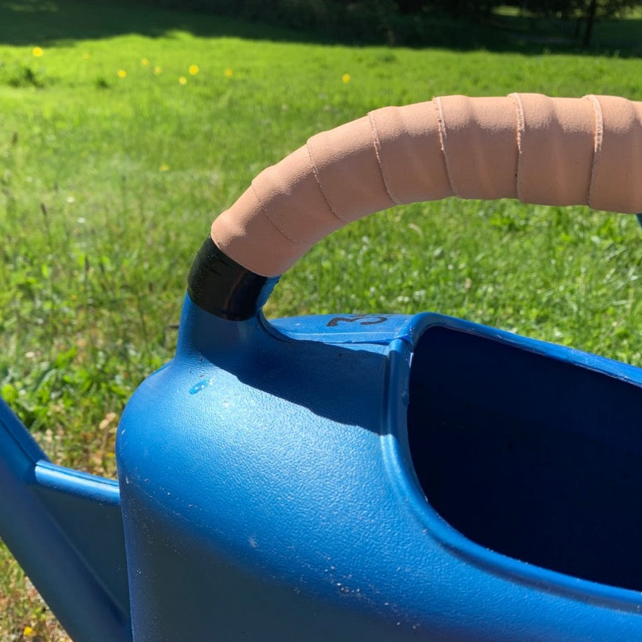 Close-up of a bright blue watering can outdoors on a sunny day. The handle of the watering can is wrapped in the Natural color Oswego leather wrap kit. 