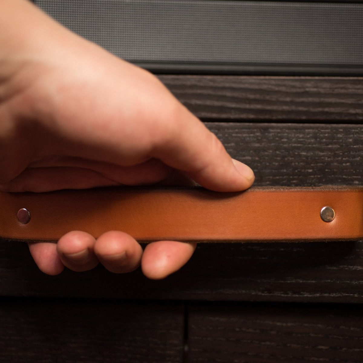 Close-up photo of a woman's hand in a large Tilikum leather handle. The handle is a honey / cognac color and the cabinet is a piece of IKEA furniture in black-brown.