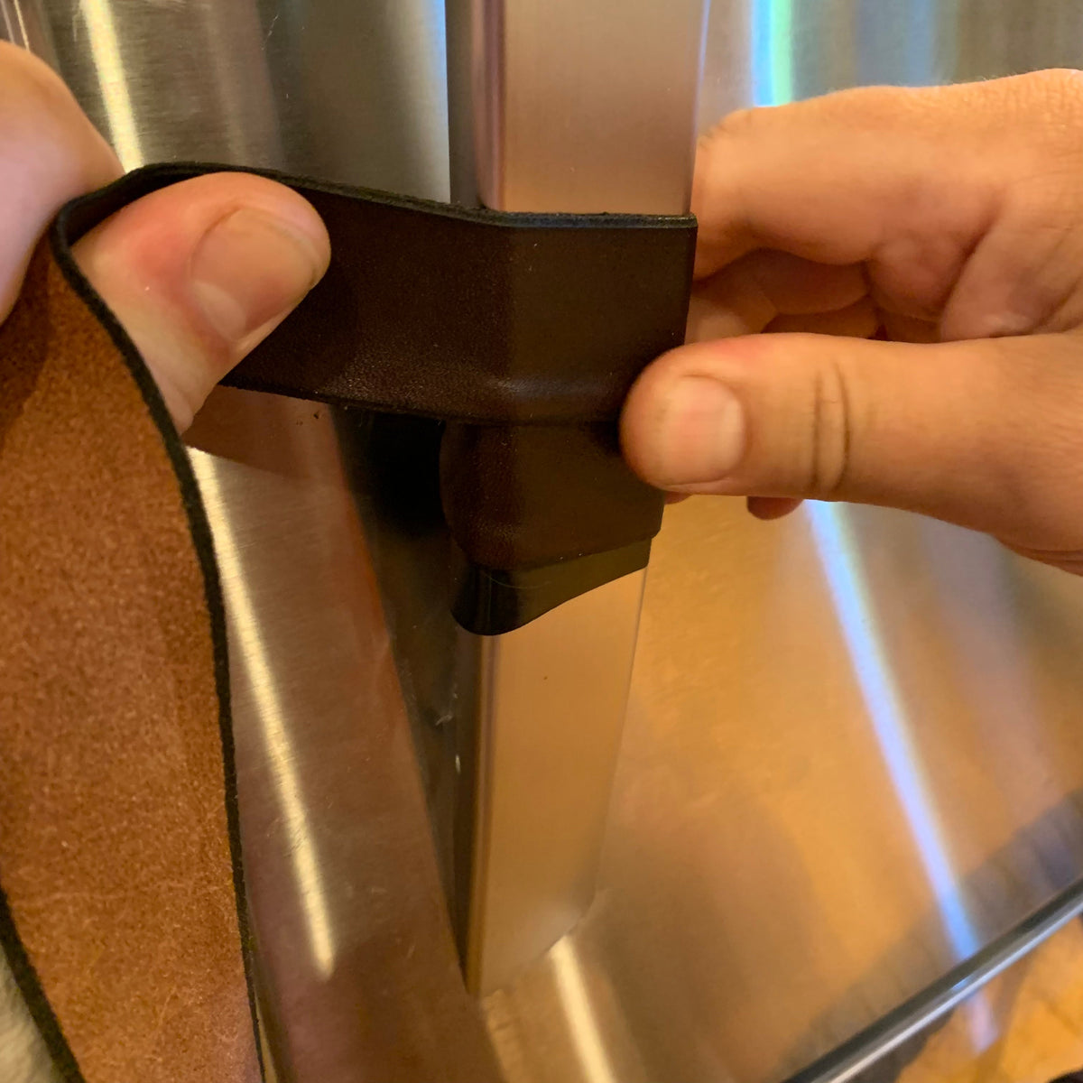 Close-up of what it looks like to wrap the Oswego leather strap around a handle: two hands are holding the leather strap as it coils around stainless steel handle clockwise. 