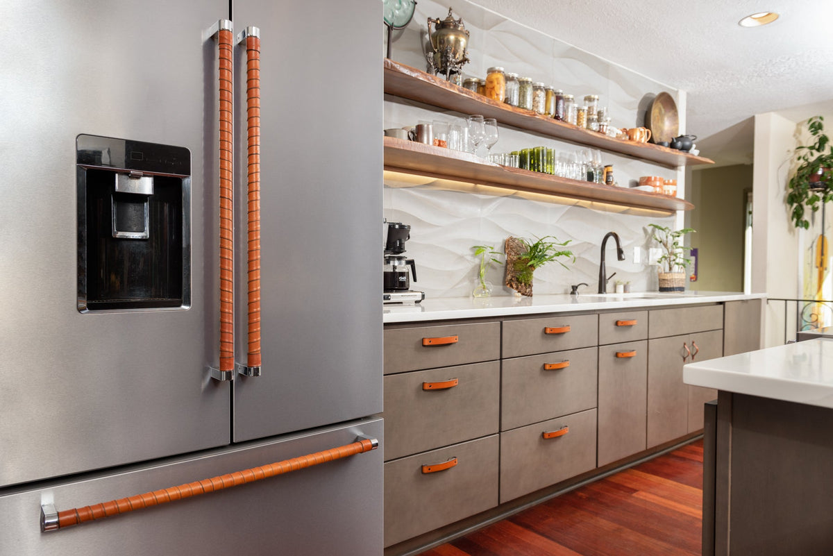 A beautiful kitchen with olive green cabinetry, custom wavy gray tile walls, and open shelving. The drawers and cabinets have Tilikum and Flanders leather drawer handles, and the refrigerator handles in the foreground are wrapped in honey Oswego leather straps. 