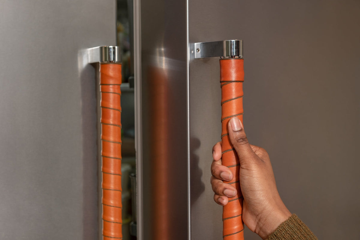Close-up of a Black woman's hand grasping a refrigerator door handle, which is wrapped in honey leather straps using the Oswego kit. The ends are covered with extra leather straps. 