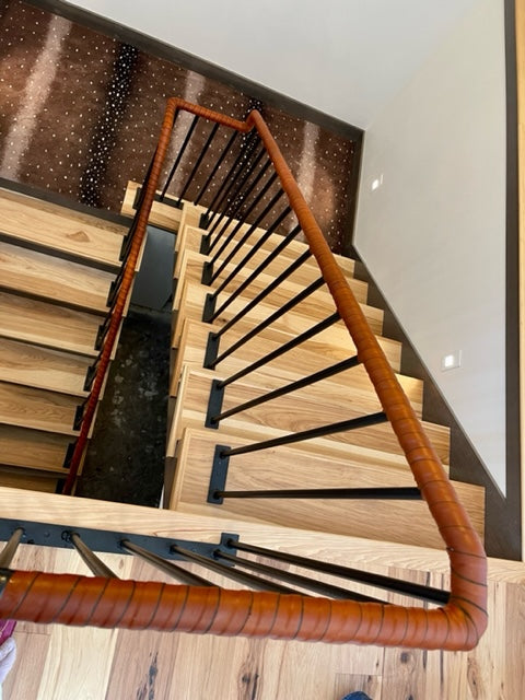 Birds-eye top-down view of a staircase landing. The stair surfaces are a light oak or birch wood and the landing is a dark starry surface. The handrail is wrapped in Oswego leather straps along the entire length down two stories. 