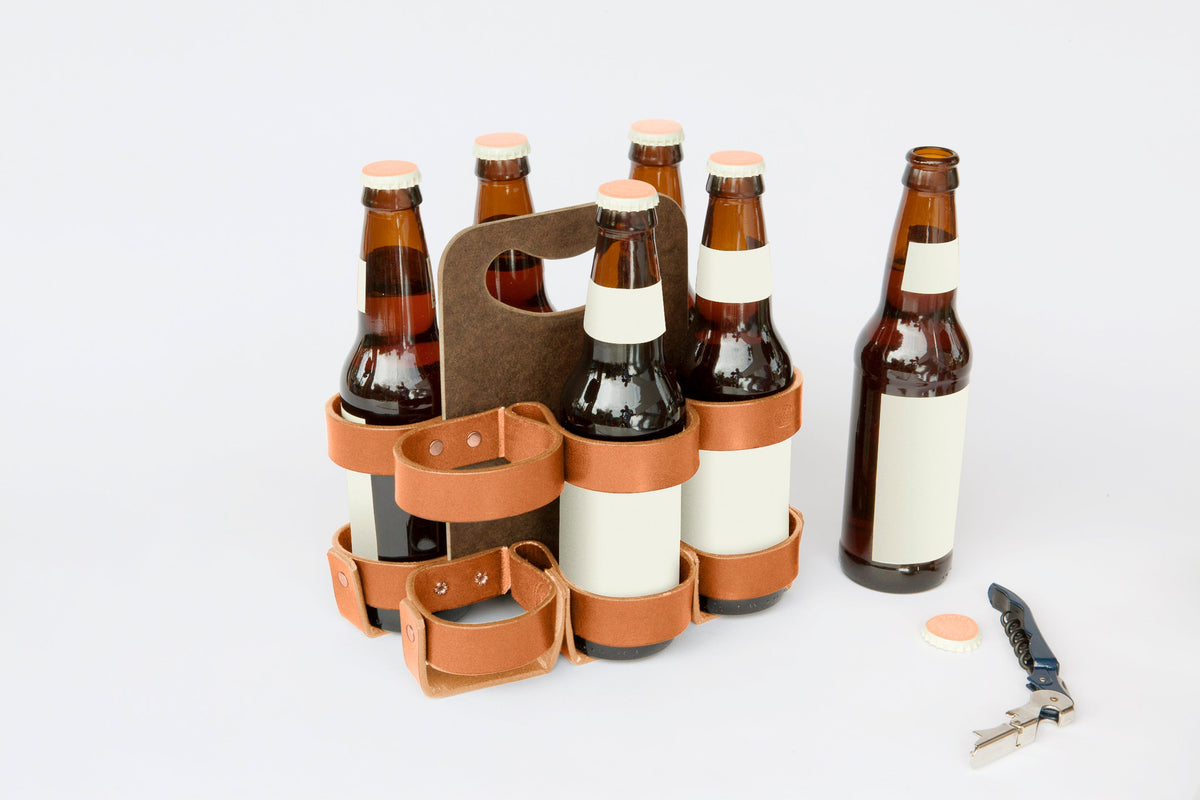 A dark brown leather and masonite six-pack carrier with six brown glass bottles of beer. A bottle opener and a bottle cap are on the table next to the carrier.