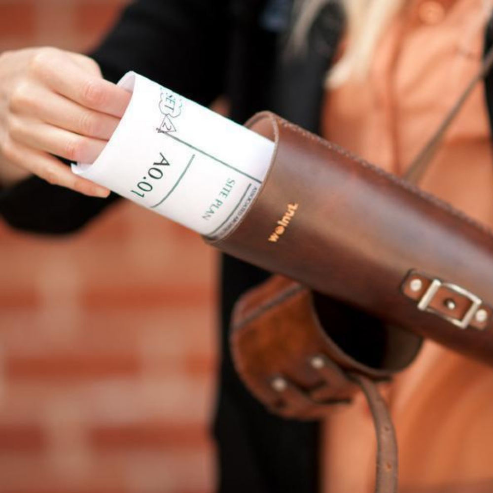 Woman pulling architectural blueprint from dark brown leather Blueprint Tube with cap removed. "walnut" is debossed on the front cover to showcase the natural leather look.
