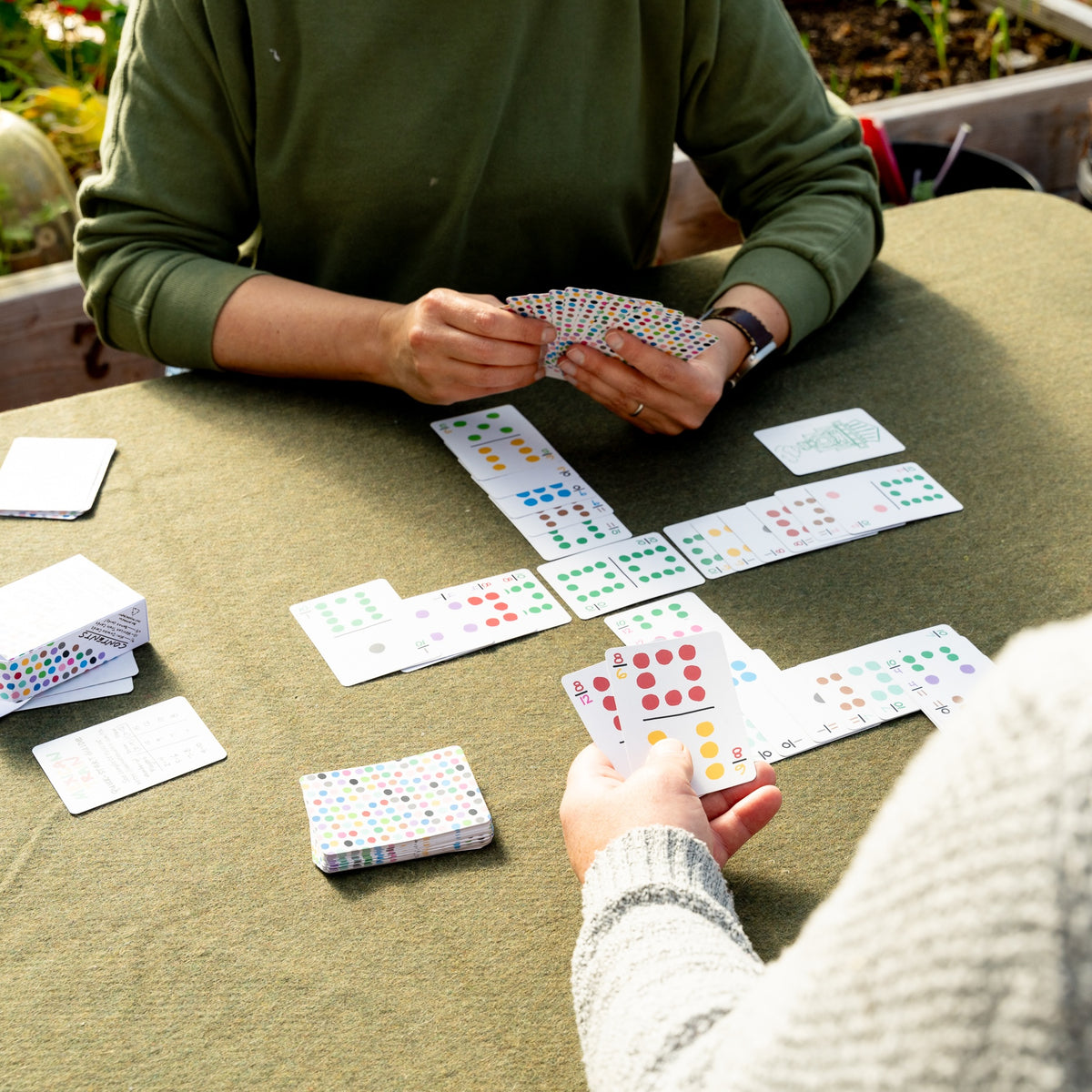 Two people playing Mexican Train outdoors in a garden on a table using standard poker playing card size travel domino playing cards. The cards are laid in trains neatly in four directions, with only the index numbers visible to save table space. The cards look comfortable to hold. 
