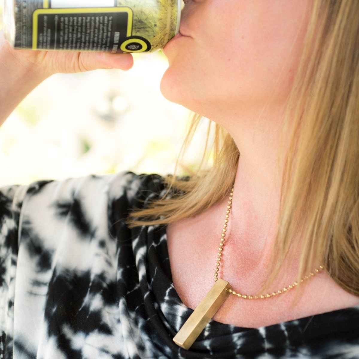 Close-up of a woman's neck and collarbone, wearing the square brass travel dice as a necklace using a longer section of brass beaded chain. 